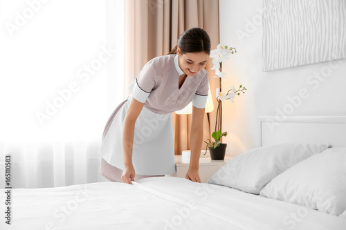 Young maid making bed in light hotel room