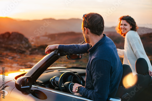A young couple have fun in a convertible at sunset, around them beautiful nature