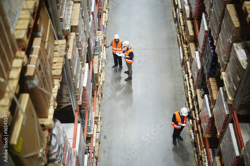 Above view of warehouse workers group in aisle between rows of tall shelves full of packed boxes and goods