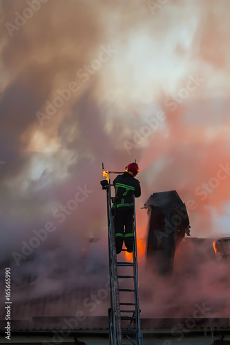 Firefighter or fireman on the ladder extinguishes burning fire flame with smoke on the apartment house roof