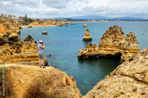 Travel photographer taking pictures of amazing landscape with digital camera while standing on a rock near the Atlantic Ocean in Lagos, Portugal 