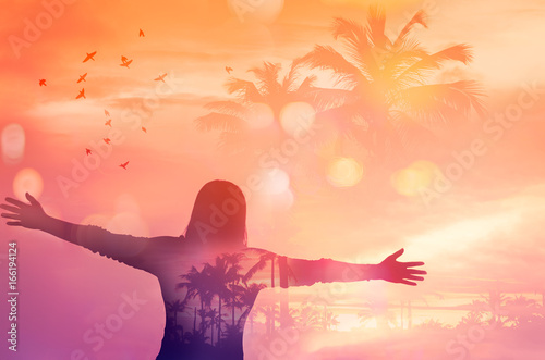 Freedom and feel good concept. Copy space of silhouette woman rising hands on sunset sky background double exposure palm tree bird fly and colorful bokeh light.