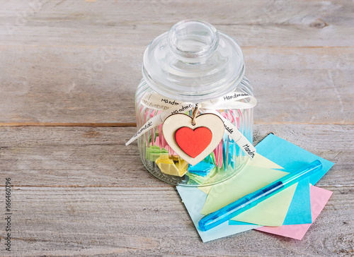 Glass jar with handmade wooden hearts decorations and ribbon near a stack of colored papers and a blue pen.