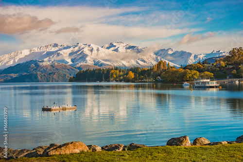 Morning snow at lakeside of Wanaka, south island, New Zealand with a view of snow mountain, colorful tree in autumn, lake and blue sky