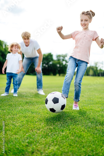 father with son looking at cute little girl kicking soccer ball on meadow