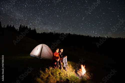 Night camping in the mountains. Romantic couple tourists have a rest at a campfire near illuminated tent under night starry sky. Low light