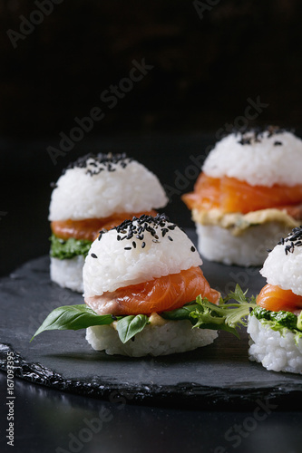 Mini rice sushi burgers with smoked salmon, green salad and sauces, black sesame served on slate stone board over black background. Modern healthy food. Close up