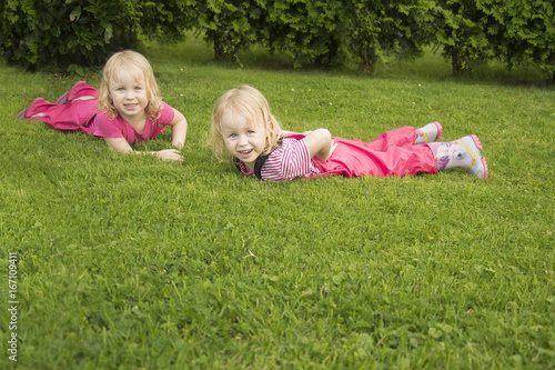 Children, two small identical girls in pink waterproof suits and rubber boots playing on a warm summer day right after the rain. Children lie directly on the damp green grass, gaily plucking it out