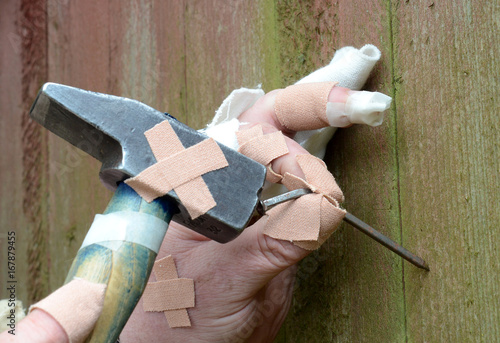 Ten thumbs, heavily "injured" homeworker tries to hammer a bent nail in a wall.