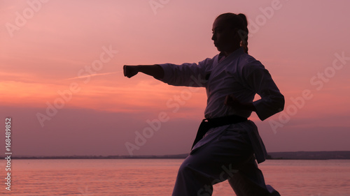 Silhouette of Caucasian female in Kimono practicing karate near large lake on sunset