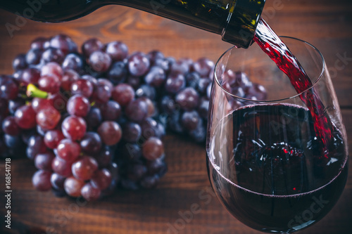 Pouring red wine into the glass with a bunch of red grapes against wooden background