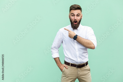 Wow! Handsome young adult man with beard in shoked. Pointing away while standing isolated on light green background