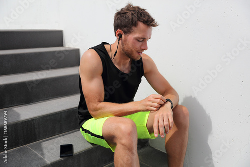 Smartwatch runner man checking progress on smart fitness sport watch during running break after hiit stairs cardio workout training. Athlete using online app on wearable device with earbuds.