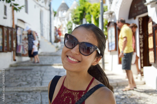 Close up of stylish young woman with sunglasses smiling in italian scenery background. Beauty woman with white perfect smile looking at camera in travel to Alberobello trulli village, Italy, Europe