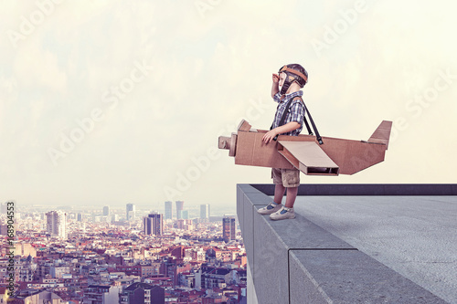  boy cardboard airplane model pilot dress looks away ledge high building. city ​​in the background. dream and imagination.