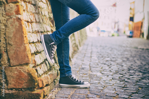A girl wearing jeans and sneakers leans on a brick wall in the historic part of town