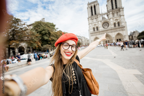 Young woman tourist in red cap making selfie photo standing in front of the famous Notre Dame cathedral in Paris
