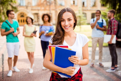 Focused shot of cheerful smart brunette caucasian girl is holding note books, smiling, standing near college building, her friends are behind, they passed tests, so cheerful and carefree!