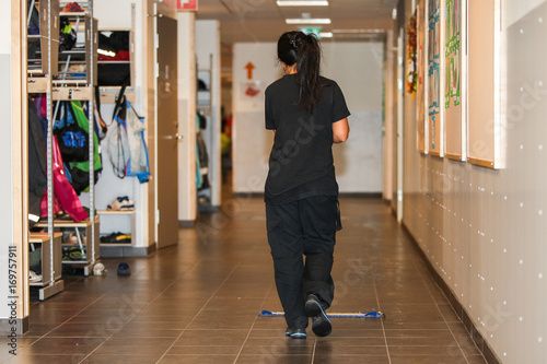 A woman, seen from behind, is sweeping and cleaning the floors in a school corridor.