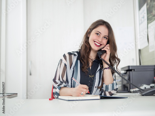 Cheerful young woman is talking on the phone while working at her office