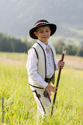 Child boy in traditional goral dress, hat and axe stands on meadow in Polish mountains