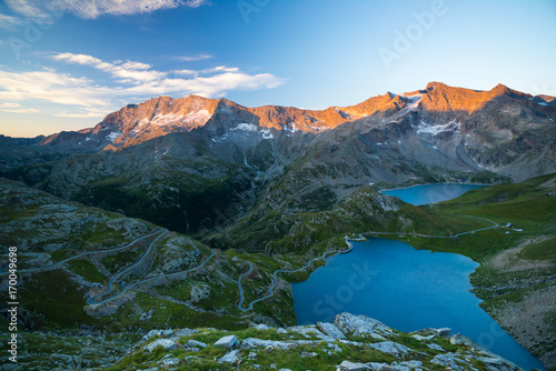High altitude alpine lake, dams and water basins in idyllic land with majestic rocky mountain peaks glowing at sunset. Road leading to mountain pass on the Alps.