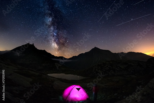 Camping with illuminated tent at high altitude on the Alps under starry sky and milky way reflected on lake. Adventure and exploration in summertime.