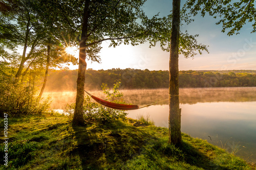 Summer camping on the lake. Empty hammock between two trees with the view of a foggy mountain lake in sunrise light. Outdoors and adventure concept.