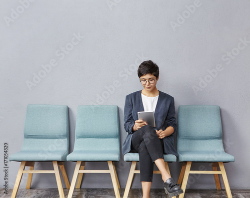 Confident successful young businesswoman with short hairstyle wearing stylish clothes and glasses using digital tablet while waiting for doctor's appointment, sitting on chair in hall at clinic