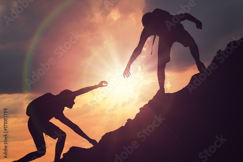 Man is giving helping hand. Silhouettes of people climbing on mountain at sunset.