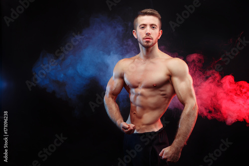 Bodybuilder man with perfect abs, shoulders,biceps, triceps and chest flexing his muscles in blue and red smoke. Strong young athlete with six pack