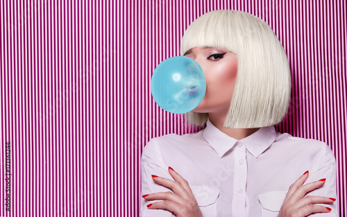 Beautiful girl in white wig blew up in pink gum bubble. A young girl in the studio on a background of a black and white vertical lines. Stylish girl wearing a white shirt.