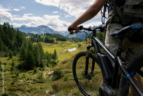 Young adult active man on mountain wearing bike helmet and backpack looking at scenic panorama holding electric bike in sunny summer day outdoor.