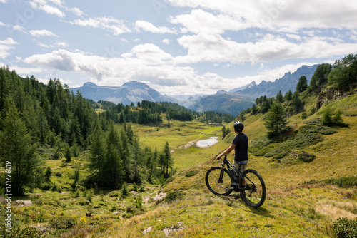 Young adult active man on mountain wearing bike helmet looking at scenic panorama holding electric bike in sunny summer day outdoor.