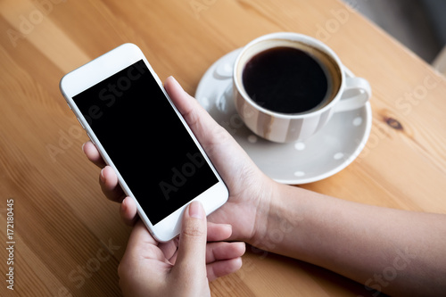 Mockup image of woman's hands holding white mobile phone with blank black screen and white coffee cups in modern loft cafe