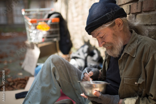 Close up view of tramp sitting by the brick wall, eating. Homeless man with his belongings living in the street.