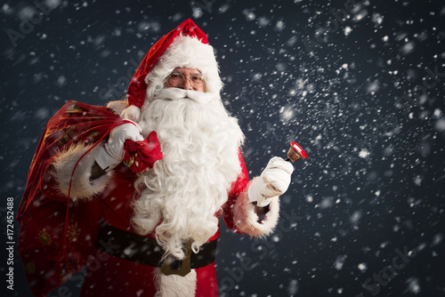 Santa Claus holding a bag with presents and ringing a bell on a dark background with snow 