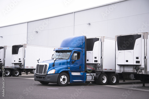 Big rig day cab semi truck and reefer trailers with refrigerator units loading cargo from warehouse building