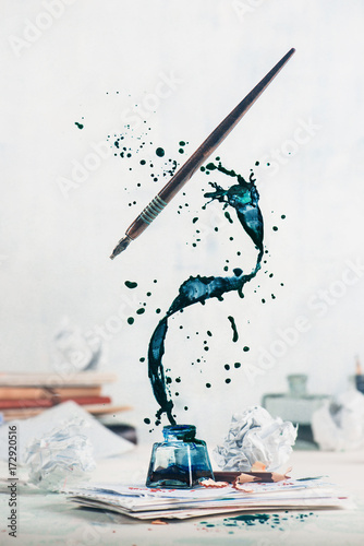 Spilled ink flying above inkwell in a spiraling splash with tiny drops and flying pen on a light background. Still life with writer workplace. Creative writing concept.