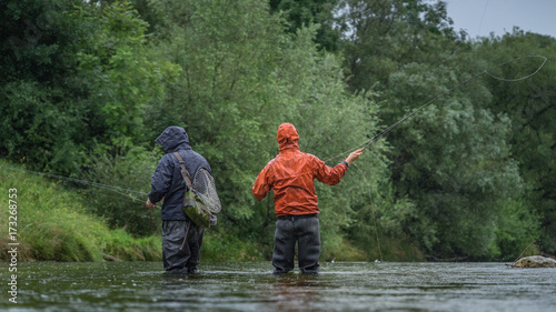 Angler mit Wathose und Watjacke im Wasser beim Angeln mit Fliegenrute bei Regen im klaren Fluss stehend und werfend