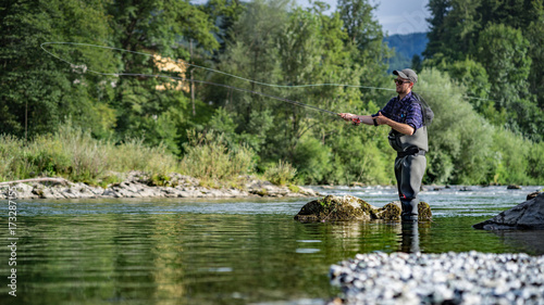 Angler mit Wathose und Fliegenrute im Wasser beim Angeln bei Sonne im klaren Fluss stehend und werfend