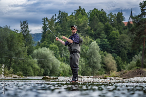 Angler mit Wathose und Fliegenrute im Wasser beim Angeln bei Sonne im klaren Fluss stehend und werfend