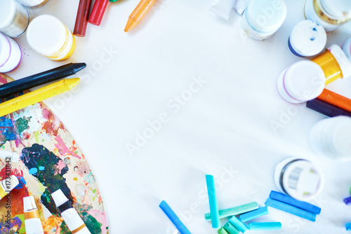 Background image of kids art supplies: colorful pencils, crayons, pastels and palette, scattered around blank white sheet of paper, copy space