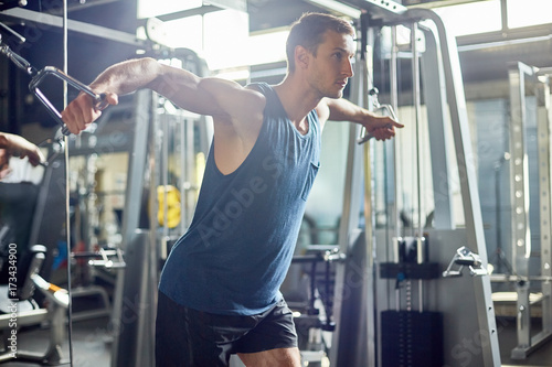 Young sporty man using cable crossover machine while having intensive workout, interior of modern gym on background