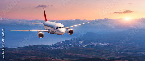 Passenger airplane. Landscape with white airplane is flying in the orange sky with clouds over mountains, sea at colorful sunset. Passenger aircraft is landing. Commercial plane. Private jet. Travel