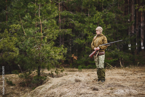 Female hunter in camouflage clothes ready to hunt, holding gun and walking in forest.