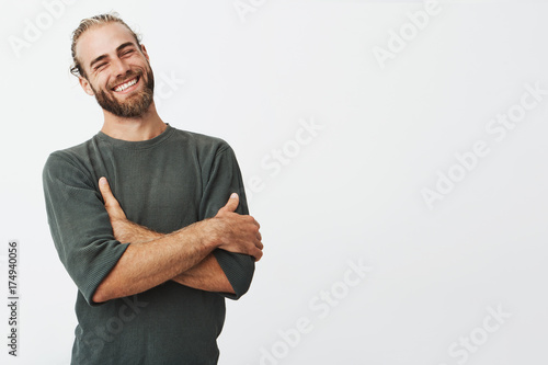 Attractive swedish man with stylish hair and beard laughs at funny story from friend with crossed hands and closed eyes.