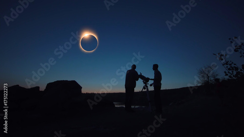 Black silhouettes of people looking through telescope on sun eclipse on shore.