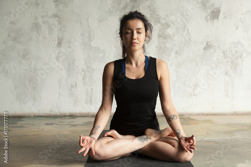 Young woman with tattoo practicing yoga, sitting in Padmasana exercise, Lotus pose, her eyes closed, working out, wearing sportswear, black shorts and top, indoor full length, studio wall background