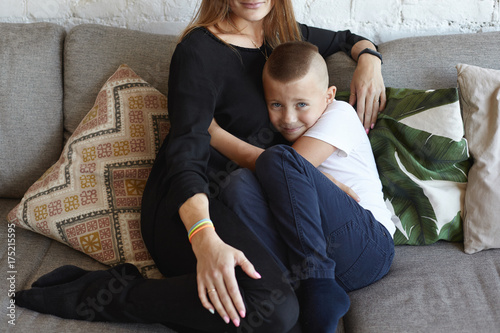 Sissy mama's boy sitting on grey couch and hugging his unrecognizable mother, seeking for her support against bullies, not able to stand up for himself, having miserable expression on his face